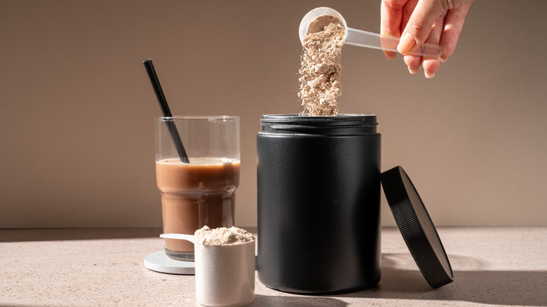 person measuring out protein powder, glass of chocolate beverage to the left and a large filled scoop in the foreground