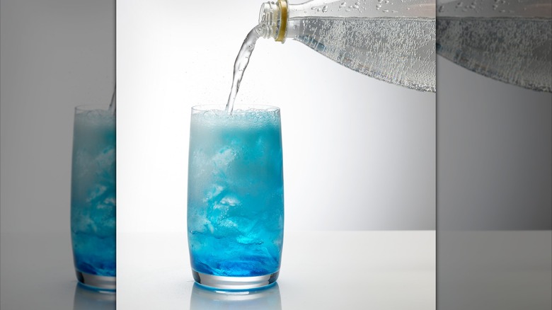 Soda being poured into blue and white bubbly mixture in a clear glass on a white background.